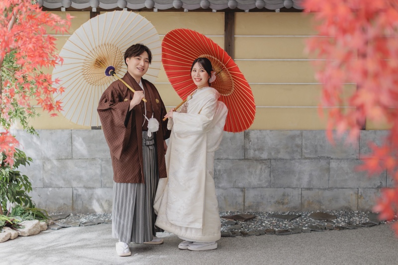 茶の羽織と白無垢で、紅葉を背景にした並び立ち
