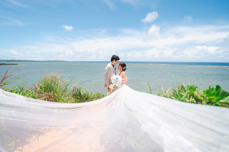 沖縄らしいどこまでも続く青い空と海をバッグに、風になびくロングベールがおふたりを優雅に包み込みます。まるで絵画のような一枚は、南国リゾートならではの開放感と、ロマンティックな雰囲気に満ち溢れています。
