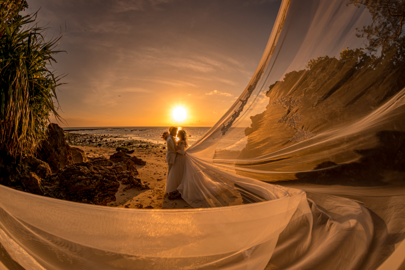 夕日のグラデーションを美しく映すために露出アンダーに、黒潰れを防ぐため逆光の被写体とヴェールにオフカメラストロボを２灯弱く当て被写体を柔らかく描写しベールの曲線を強調するために超広角で撮影しました。