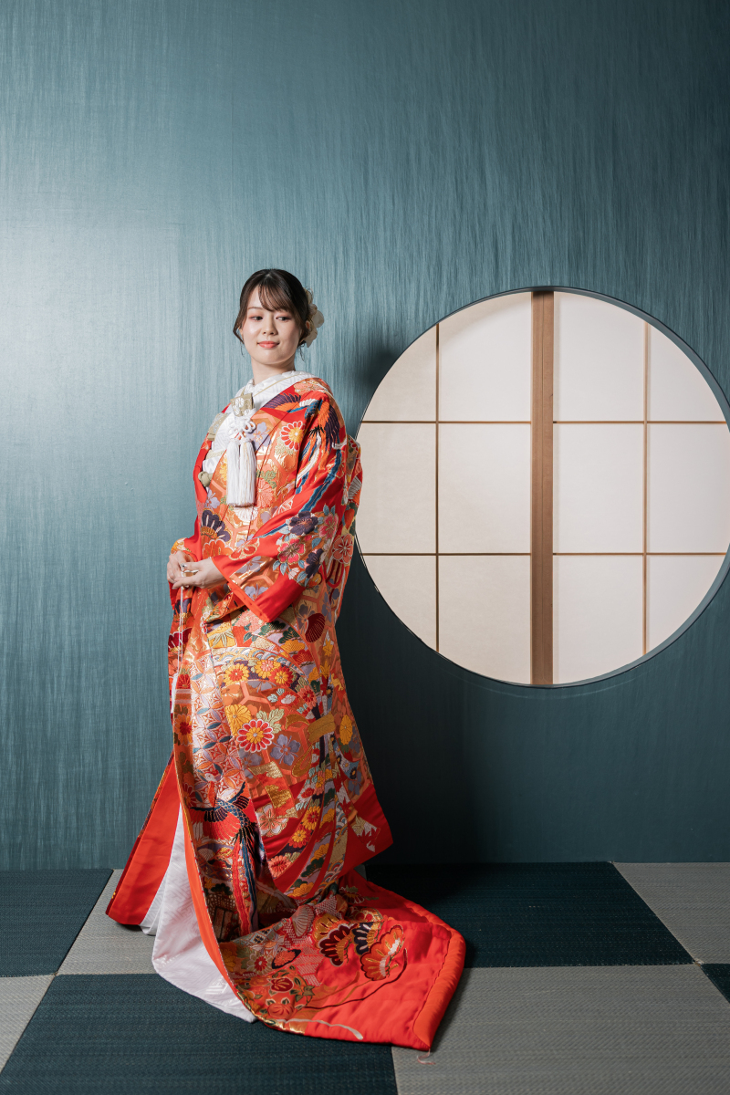 丸窓の前で斜め向きに立つ色打掛の新婦様