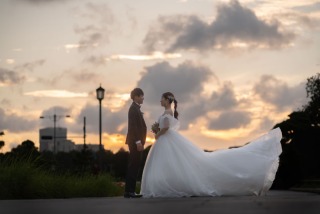 オレンジ色に染まる夕暮れの空を背景に、微笑み合いながら見つめ合う新郎新婦。風に大きく舞い上がる純白のウェディングドレスが、まるで映画のワンシーンのようにドラマチックでロマンチックな一枚です。