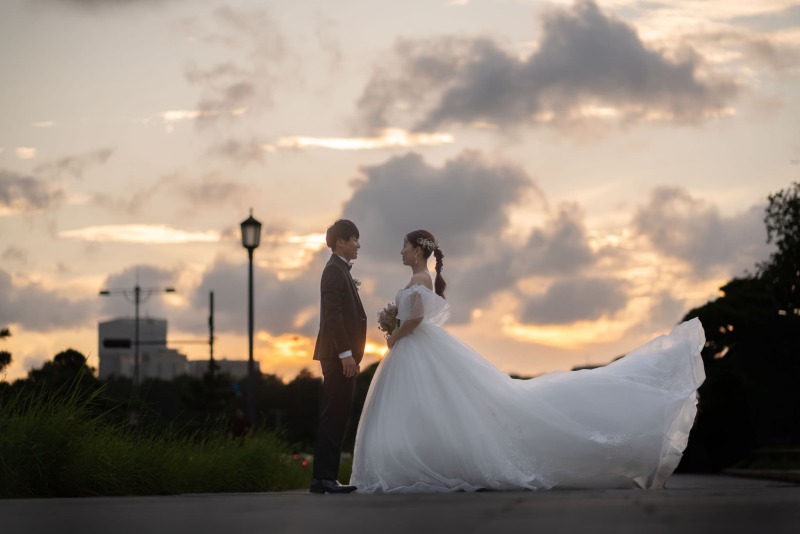 オレンジ色に染まる夕暮れの空を背景に、微笑み合いながら見つめ合う新郎新婦。風に大きく舞い上がる純白のウェディングドレスが、まるで映画のワンシーンのようにドラマチックでロマンチックな一枚です。